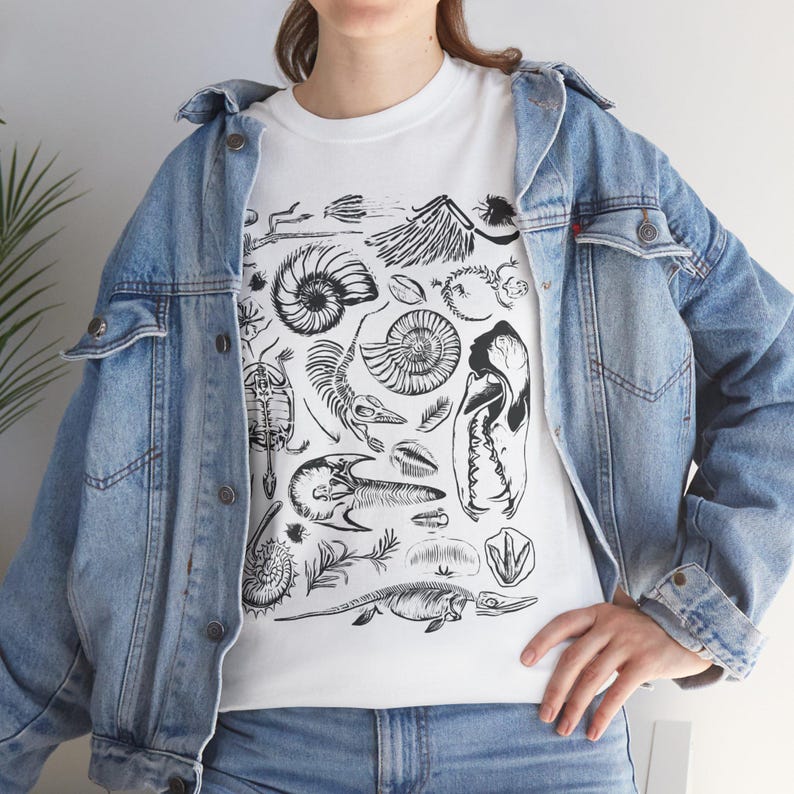 May include: A white t-shirt featuring a black ink graphic print of various fossils, including ammonites, a turtle shell, a skull, and prehistoric skeletal remains, worn under a light blue denim jacket with metal buttons and chest pockets.