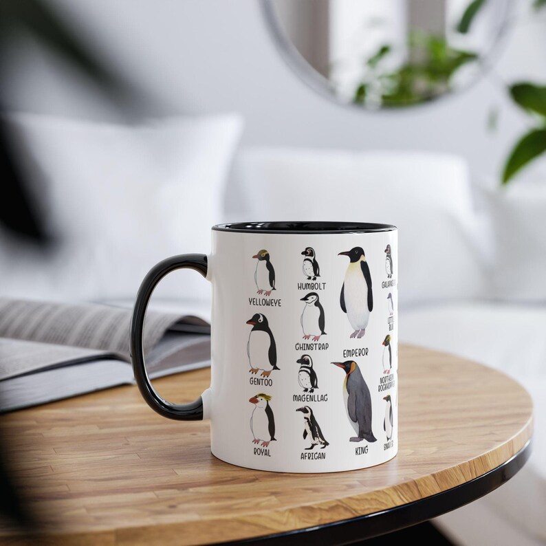 May include: White ceramic mug with a black interior, handle, and rim. The mug features illustrations of various penguin species with their names. The mug is on a wooden table.