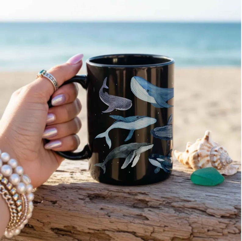 May include: Black ceramic mug featuring watercolor illustrations of various whale species. The mug has a black handle and a glossy finish. The whales are depicted in shades of blue and gray, with white accents. The mug is held near a beach setting.