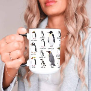 May include: White ceramic mug featuring illustrations of various penguin species, including Yelloweye, Humboldt, and Emperor penguins. Each penguin is labeled with its name. The mug has a black handle and rim, and is held by a person.