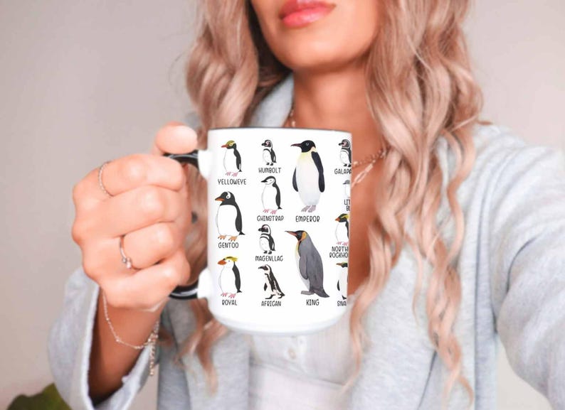 May include: White ceramic mug featuring illustrations of various penguin species, including Yelloweye, Humboldt, and Emperor penguins. Each penguin is labeled with its name. The mug has a black handle and rim, and is held by a person.