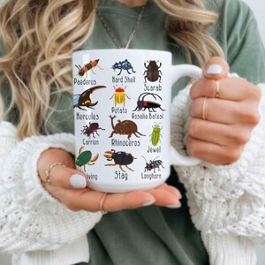 K&ouml;nnte beinhalten: Wei&szlig;e Keramiktasse mit farbenfrohen Illustrationen verschiedener Insekten, jeweils mit Namen versehen. Die Tasse hat einen gro&szlig;en Henkel und wird von einer Person gehalten, die einen wei&szlig;en Pullover tr&auml;gt.
