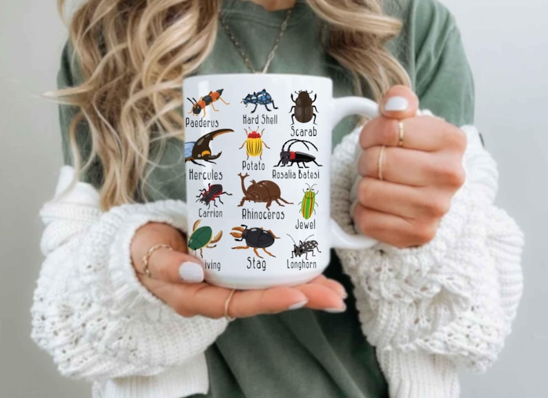 K&ouml;nnte beinhalten: Wei&szlig;e Keramiktasse mit farbenfrohen Illustrationen verschiedener Insekten, jeweils mit Namen versehen. Die Tasse hat einen gro&szlig;en Henkel und wird von einer Person gehalten, die einen wei&szlig;en Pullover tr&auml;gt.