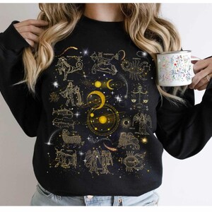 May include: Black sweatshirt with a gold zodiac constellation print on a dark blue background. The constellations include Aries, Taurus, Gemini, Cancer, Leo, Virgo, Libra, Scorpio, Sagittarius, Capricorn, Aquarius, and Pisces.