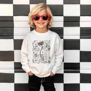 Op de afbeelding: Een wit sweatshirt met lange mouwen en een zwarte inktachtige grafische print van diverse skeletresten, fossielen en insecten op de borst, gecombineerd met een zonnebril met rood montuur en een zwarte broek.