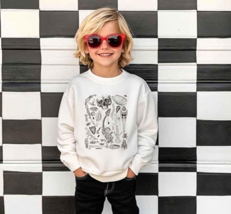 Op de afbeelding: Een wit sweatshirt met lange mouwen en een zwarte inktachtige grafische print van diverse skeletresten, fossielen en insecten op de borst, gecombineerd met een zonnebril met rood montuur en een zwarte broek.