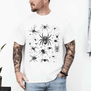 May include: A white short-sleeved t-shirt featuring a central graphic print of various black spiders in different sizes and species. The design displays a collection of detailed arachnid illustrations arranged in a cluster on the front of the shirt.