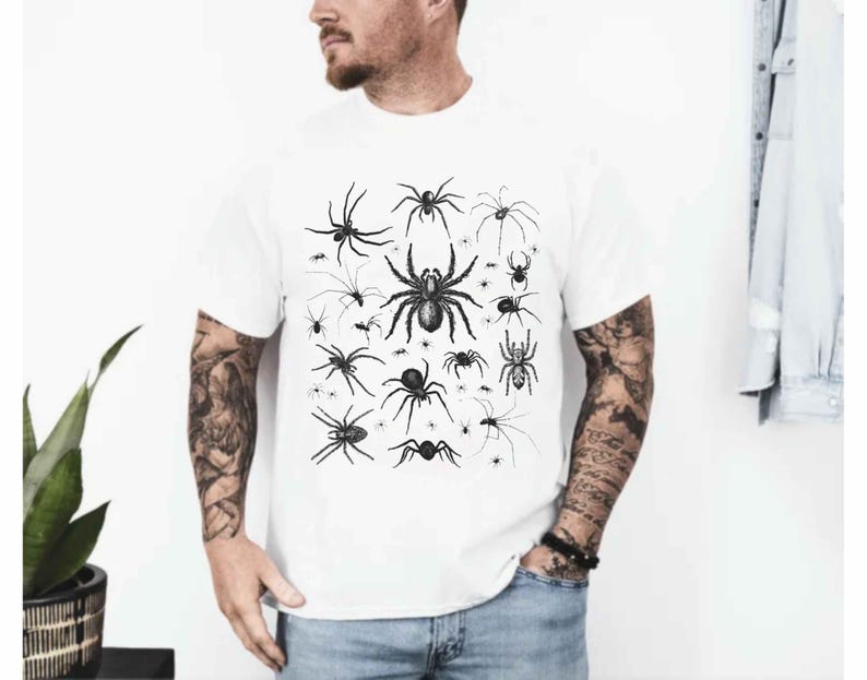 May include: A white short-sleeved t-shirt featuring a central graphic print of various black spiders in different sizes and species. The design displays a collection of detailed arachnid illustrations arranged in a cluster on the front of the shirt.