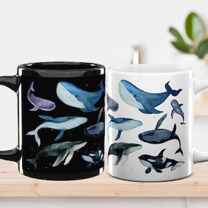 May include: Two ceramic mugs, one black and one white, each decorated with watercolour illustrations of different whale species in shades of blue and purple. The mugs are on a light wooden surface.
