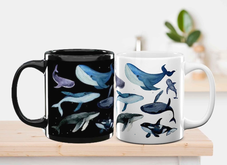 May include: Two ceramic mugs, one black and one white, each decorated with watercolour illustrations of different whale species in shades of blue and purple. The mugs are on a light wooden surface.