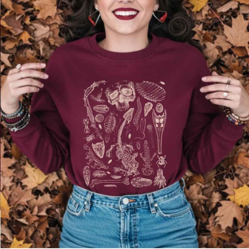 May include: Burgundy-colored sweatshirt featuring a detailed cream-colored anatomical illustration of bones, insects, and a skull. The design is centered on the front of the long-sleeved crew neck top. The model is wearing blue jeans.