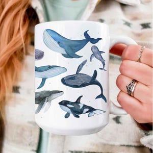 May include: White ceramic mug with watercolour illustrations of different whales in shades of blue. The mug has a large handle and a smooth, glossy finish. The whale designs include a humpback, orca, and other whale species.