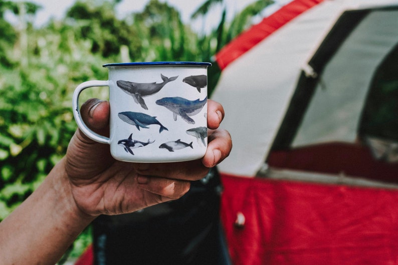 Peut inclure: Une tasse en &eacute;mail blanc avec un bord bleu, orn&eacute;e d'un motif de diff&eacute;rentes baleines dans des tons de gris et de noir. La tasse est tenue par une main devant une tente rouge et blanche.