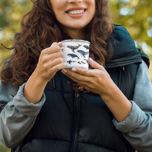 Peut inclure: Une tasse en &eacute;mail blanche avec un motif de baleine noir et bleu. La tasse est tenue par une personne portant un gilet noir et une chemise grise.