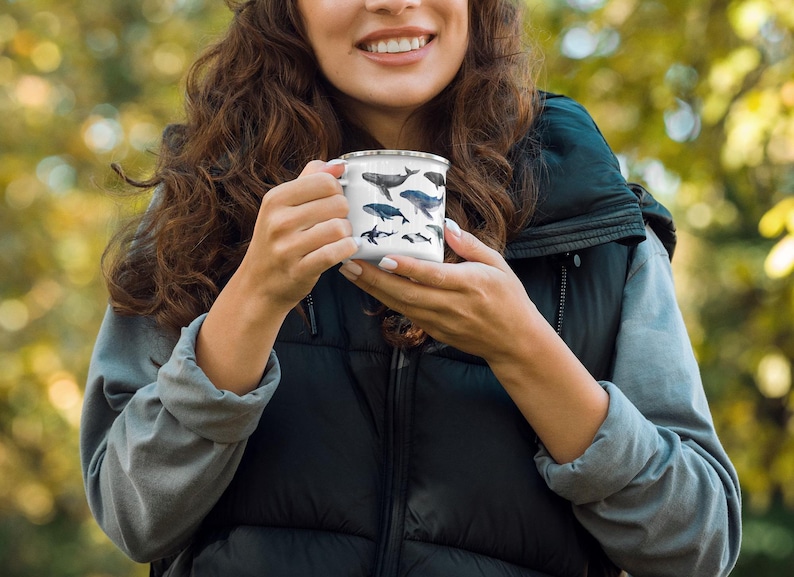 Peut inclure: Une tasse en &eacute;mail blanche avec un motif de baleine noir et bleu. La tasse est tenue par une personne portant un gilet noir et une chemise grise.