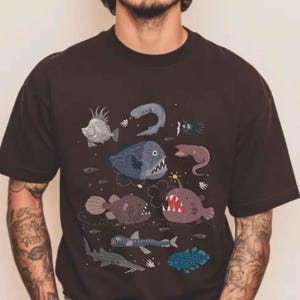 May include: A dark brown short-sleeved t-shirt featuring a graphic print of various deep-sea creatures, including an anglerfish with a glowing lure, a shark, and several other marine animals in shades of blue, grey, and purple across the chest.