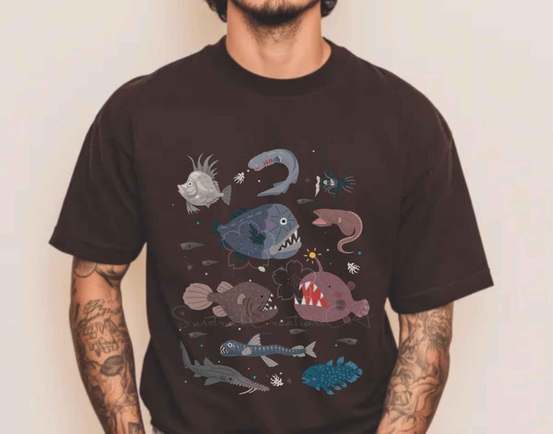 May include: A dark brown short-sleeved t-shirt featuring a graphic print of various deep-sea creatures, including an anglerfish with a glowing lure, a shark, and several other marine animals in shades of blue, grey, and purple across the chest.