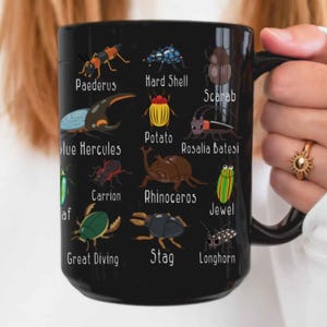 K&ouml;nnte beinhalten: Schwarze Keramik-Tasse mit farbenfrohen Illustrationen verschiedener K&auml;fer und ihren Namen. Die Tasse zeigt K&auml;fer wie den Blauen Herkules und den Hirschk&auml;fer. Ein einzigartiges Geschenk f&uuml;r Entomologie-Enthusiasten.