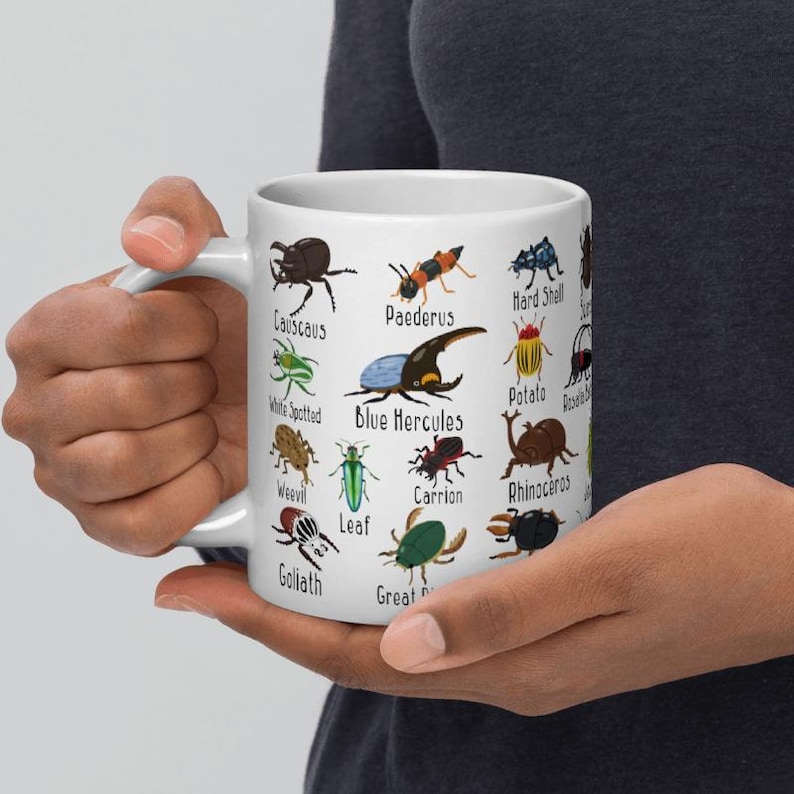 K&ouml;nnte beinhalten: Wei&szlig;e Keramiktasse mit farbenfrohen Illustrationen verschiedener Insekten, die jeweils mit ihrem Namen beschriftet sind. Die Tasse wird von einer Person gehalten, die die detaillierten Insektenmuster und den Text, einschlie&szlig;lich "Blue Hercules" und "Goliath", zeigt.