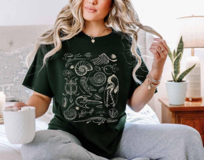 May include: A dark green short-sleeve t-shirt featuring a white graphic print of various fossils, including ammonites, a turtle shell, and skeletal remains, arranged in a collage pattern across the center of the chest area.