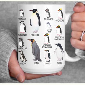 May include: White ceramic mug featuring illustrations of various penguin species, including Humboldt, Emperor, and Macaroni penguins. Each penguin is labeled with its name. The mug is held by a person.
