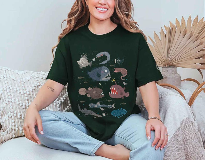 May include: A dark green short-sleeved t-shirt featuring a graphic print of various deep-sea creatures, including an anglerfish, a shark, and other marine life, arranged in a scattered pattern across the centre of the chest area.