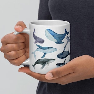 May include: White ceramic mug decorated with watercolour illustrations of different whale species in shades of blue and grey. The mug has a standard handle and a glossy finish, ideal for hot drinks.