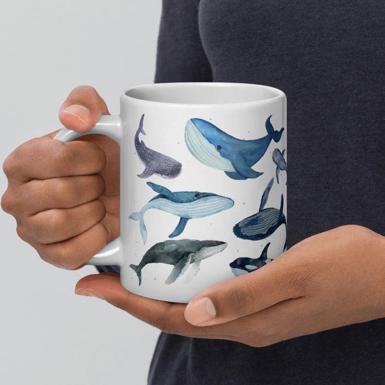 May include: White ceramic mug decorated with watercolour illustrations of different whale species in shades of blue and grey. The mug has a standard handle and a glossy finish, ideal for hot drinks.
