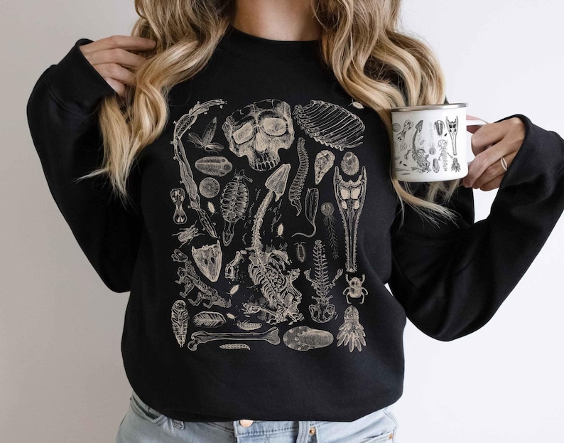 Op de afbeelding: Een zwart sweatshirt met lange mouwen en een gedetailleerde beige grafische print van diverse biologische specimens, waaronder een schedel, skeletstructuren, insecten en fossielen in een rasterpatroon op de voorzijde.