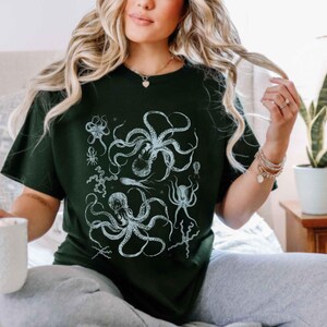 May include: A dark green short-sleeved t-shirt featuring a detailed white graphic print of multiple octopuses and various sea creatures arranged in a cluster on the front center of the garment.