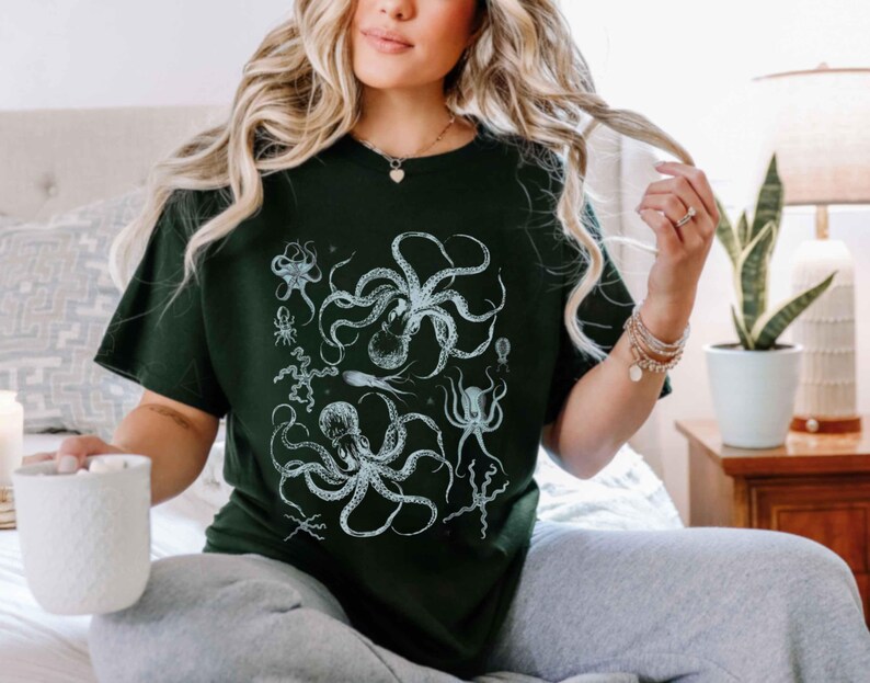May include: A dark green short-sleeved t-shirt featuring a detailed white graphic print of multiple octopuses and various sea creatures arranged in a cluster on the front center of the garment.