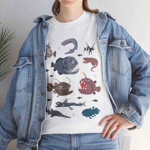 May include: A white t-shirt featuring a graphic print of various deep-sea creatures, including an anglerfish, a sawshark, and an eel, layered under a blue denim jacket. The design displays multiple marine animals in shades of blue, brown, and grey.