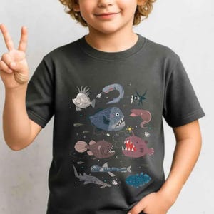 May include: A dark grey short-sleeved t-shirt featuring a graphic print of various deep-sea creatures, including an anglerfish with a glowing lure, a fangtooth fish, and other marine life illustrated in shades of blue, red, and brown.