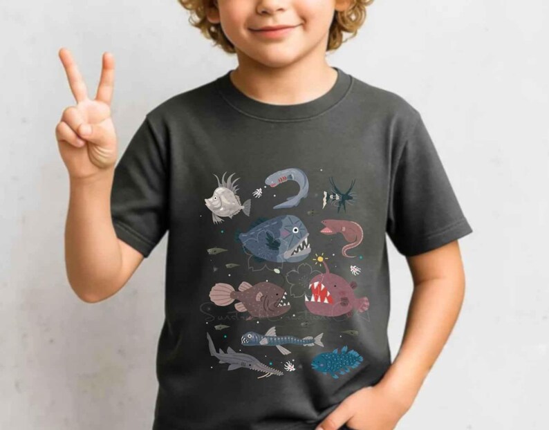 May include: A dark grey short-sleeved t-shirt featuring a graphic print of various deep-sea creatures, including an anglerfish with a glowing lure, a fangtooth fish, and other marine life illustrated in shades of blue, red, and brown.