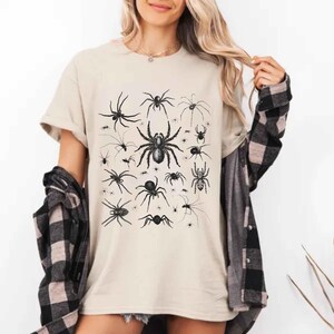 May include: A beige short-sleeved t-shirt featuring a black graphic print of various spider species in different sizes and orientations. The shirt is worn with a black and beige plaid long-sleeved button-down shirt layered over the top.