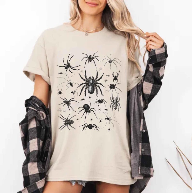 May include: A beige short-sleeved t-shirt featuring a black graphic print of various spider species in different sizes and orientations. The shirt is worn with a black and beige plaid long-sleeved button-down shirt layered over the top.