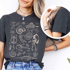 May include: A dark charcoal gray t-shirt featuring a detailed cream-colored graphic print of various prehistoric fossils, including ammonites, a turtle skeleton, and other skeletal remains arranged in a collage across the front of the garment.
