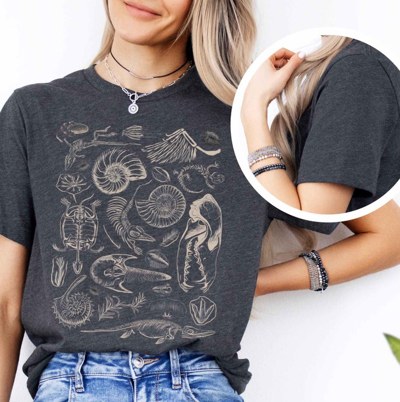 May include: A dark charcoal gray t-shirt featuring a detailed cream-colored graphic print of various prehistoric fossils, including ammonites, a turtle skeleton, and other skeletal remains arranged in a collage across the front of the garment.