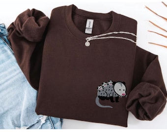 May include: A dark brown crewneck sweatshirt featuring an embroidered graphic of a gray opossum with three small babies on its back. The opossum has black paws, a pink nose, and a long gray tail. The sweatshirt has a ribbed collar and cuffs.
