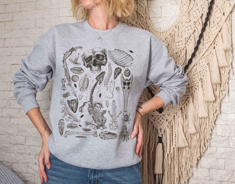 Op de afbeelding: Een lichtgrijs sweatshirt met ronde hals voorzien van een gedetailleerde zwarte inktillustratie van diverse biologische specimens, waaronder een menselijke schedel, skeletresten, insectenanatomie en botanische elementen.