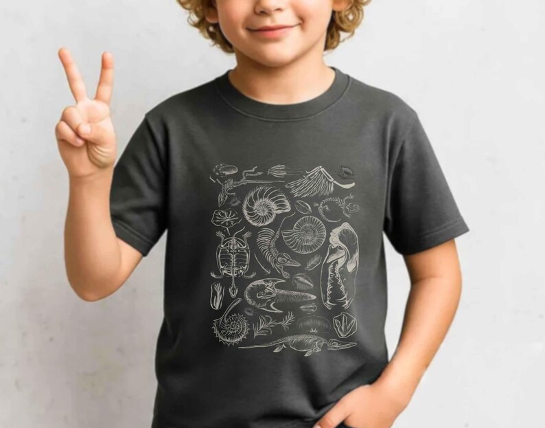 May include: A dark gray short-sleeve t-shirt featuring a white graphic print of various prehistoric fossils, including ammonites, skeletal remains, and marine life silhouettes arranged in a rectangular pattern on the front of the garment.