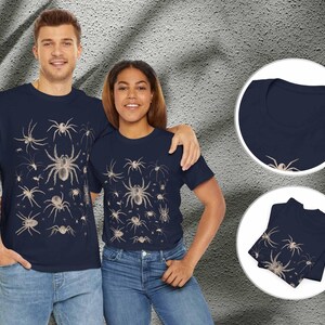 May include: A navy blue short-sleeved t-shirt featuring a repeating pattern of various spider illustrations in a light beige colour. The design is displayed on the front of the shirt, with additional circular insets showing the neckline and folded fabric.