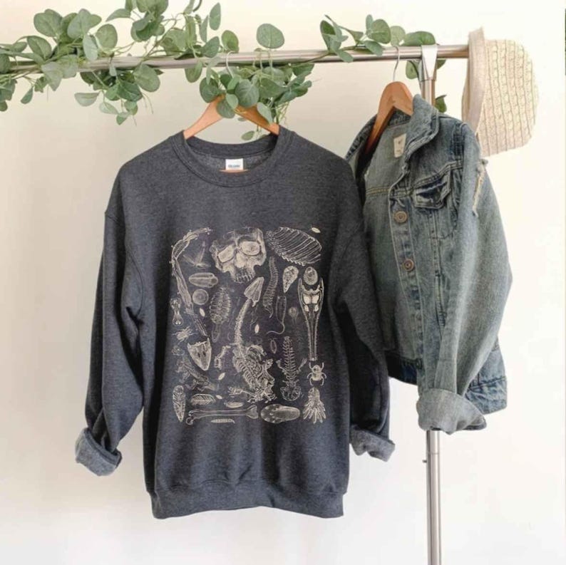 Op de afbeelding: Een donkergrijze sweater met ronde hals en een witte grafische print van diverse gefossiliseerde skeletresten, waaronder een schedel, ruggengraat en insect, gepresenteerd naast een blauw spijkerjack op een kledingrek.