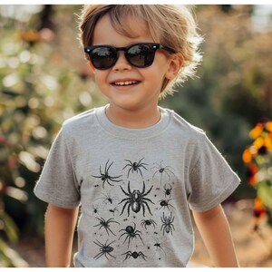 May include: A light grey short-sleeved t-shirt featuring a black graphic print of various spider species in different sizes and orientations across the front chest area. The shirt has a crew neckline and a standard fit, showcasing detailed illustrations.
