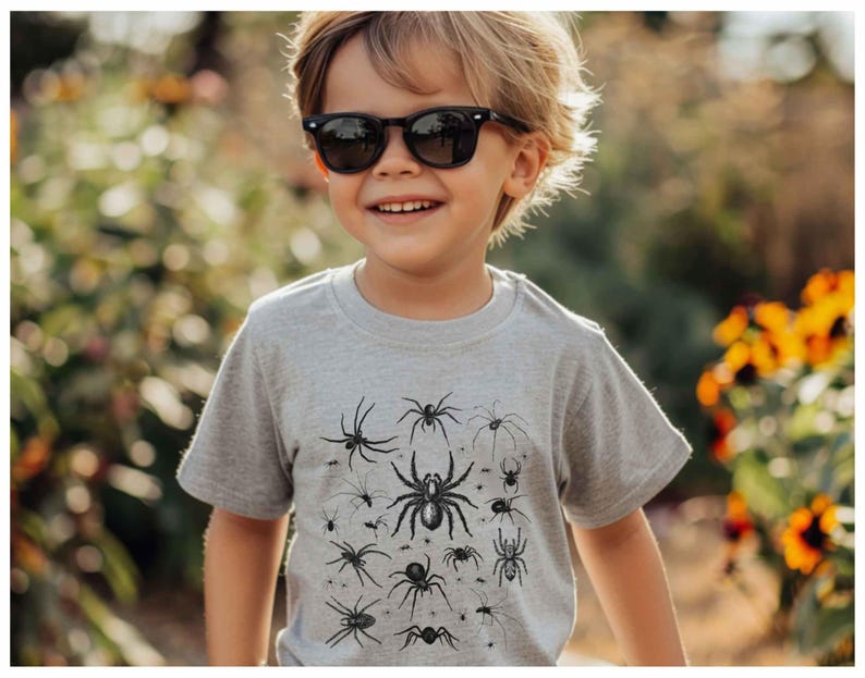 May include: A light grey short-sleeved t-shirt featuring a black graphic print of various spider species in different sizes and orientations across the front chest area. The shirt has a crew neckline and a standard fit, showcasing detailed illustrations.