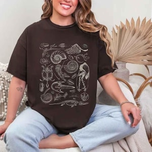 May include: A dark brown short-sleeved t-shirt featuring a white graphic print of various fossils, including ammonites, skeletal remains, and prehistoric creatures arranged in a central rectangular pattern on the front of the garment.