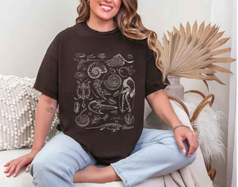 May include: A dark brown short-sleeved t-shirt featuring a white graphic print of various fossils, including ammonites, skeletal remains, and prehistoric creatures arranged in a central rectangular pattern on the front of the garment.