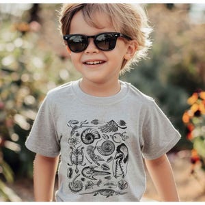 May include: A light gray short-sleeved t-shirt featuring a black graphic print of various fossils, including ammonites, skeletal remains, and prehistoric creatures, worn with black-framed sunglasses.