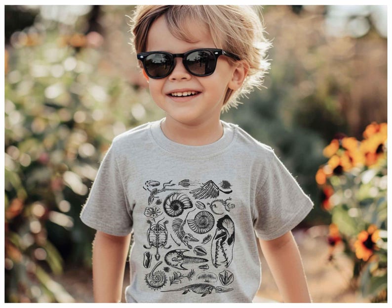May include: A light gray short-sleeved t-shirt featuring a black graphic print of various fossils, including ammonites, skeletal remains, and prehistoric creatures, worn with black-framed sunglasses.