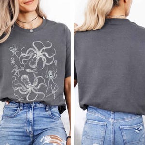 May include: A charcoal gray short-sleeve t-shirt featuring a white graphic print of multiple octopuses and sea creatures on the front. The back of the t-shirt is plain charcoal gray. The shirt is paired with blue denim jeans with distressed details.